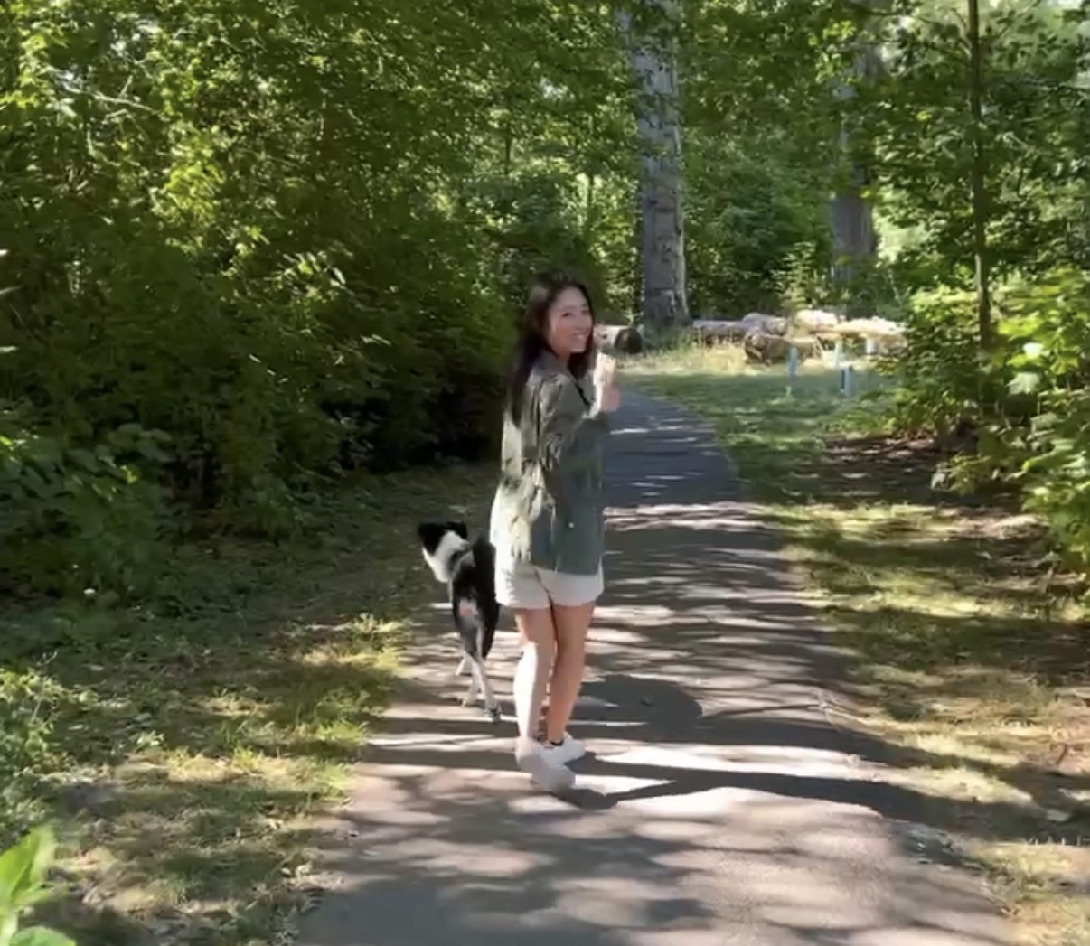 Elaine Oyang standing on a sunlit wooded path with a black and white dog