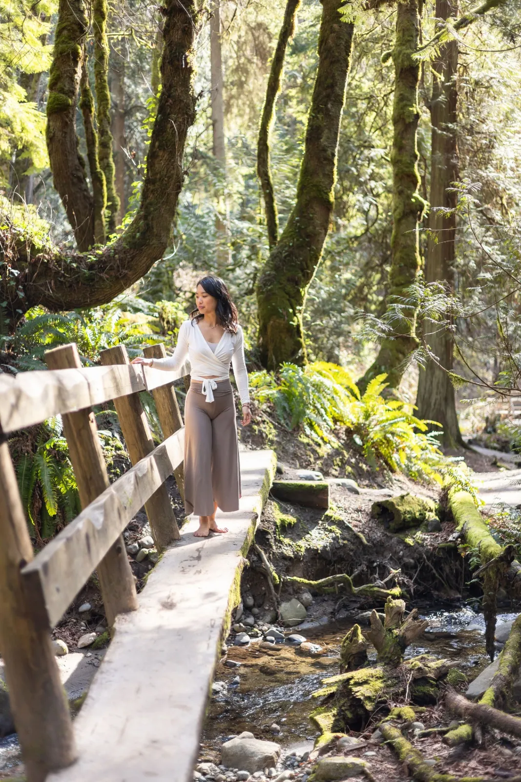 Elaine walking barefoot across a small wooden footbridge through a sunlit, mossy Pacific Northwest forest.