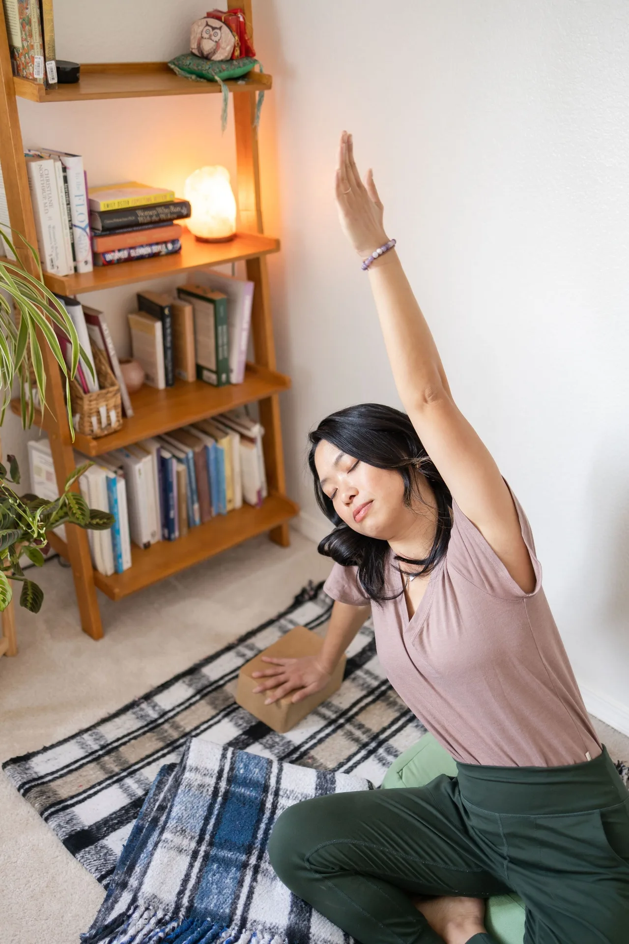 Elaine kneeling on a woven plaid rug in a softly lit home practice space, eyes closed, right arm sweeping overhead and left hand resting on a cork yoga block in a supported side stretch that opens the ribs and side body.